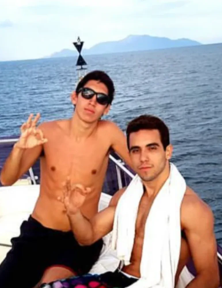 Two friends on a boat making OK gestures with the open sea and a mountain behind