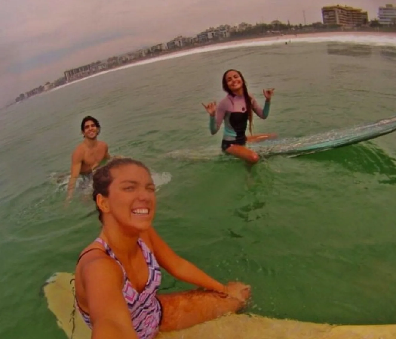 Friends smiling on surfboards in the green water, one making a shaka sign