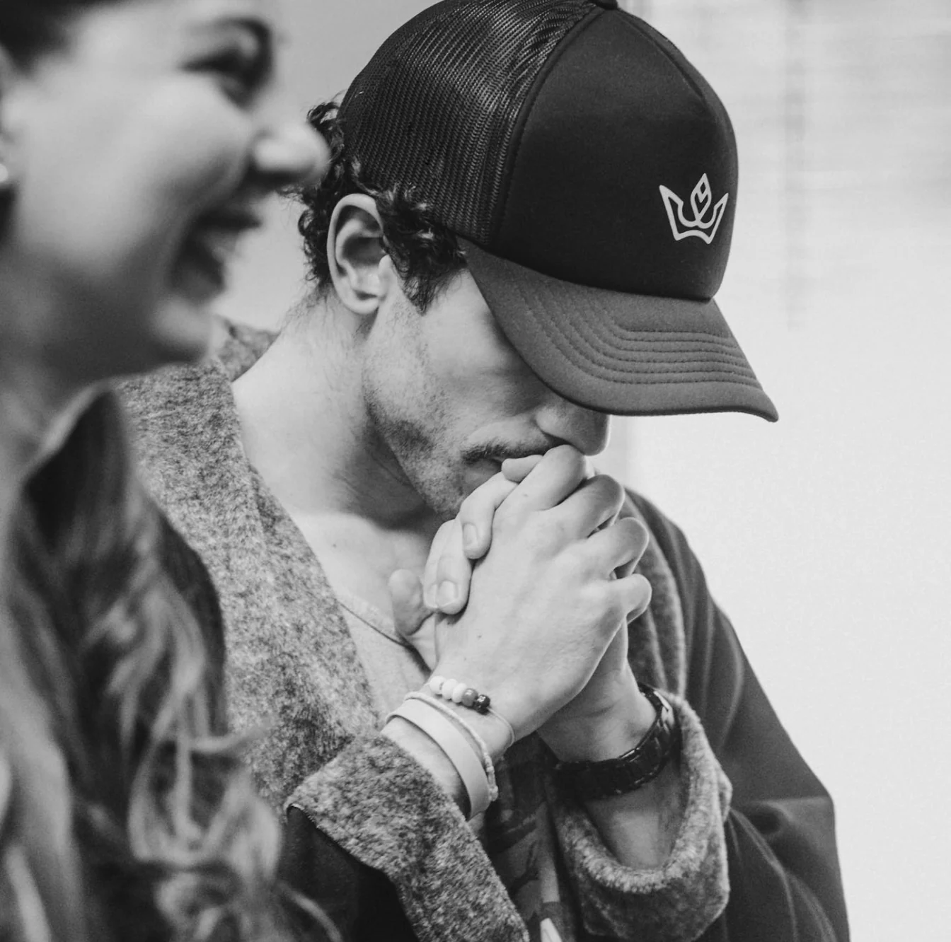 Black and white portrait of Raphael in a cap with hands together, a woman laughing in the foreground