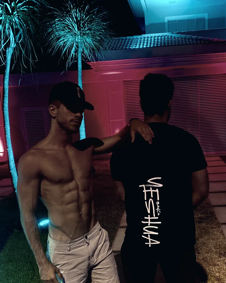 Raphael with a friend at night under neon-lit palm trees, the friend's black shirt reads Yeshua