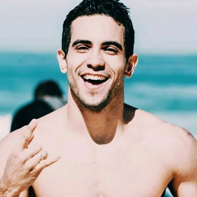 Raphael smiling big with a shaka sign in front of the sea