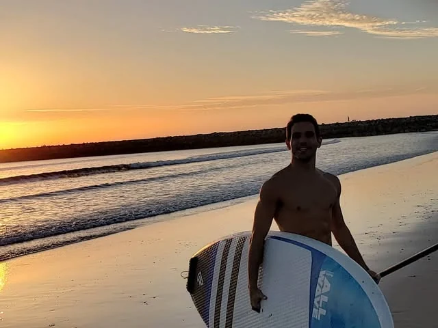 Raphael carrying a paddle board along the beach at sunset