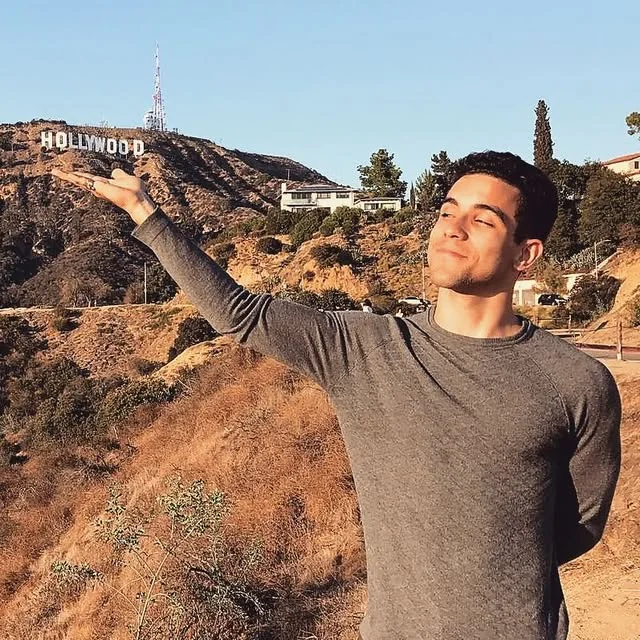 Raphael outdoors in a gray long-sleeve shirt, arm stretched out, with dry hills behind him